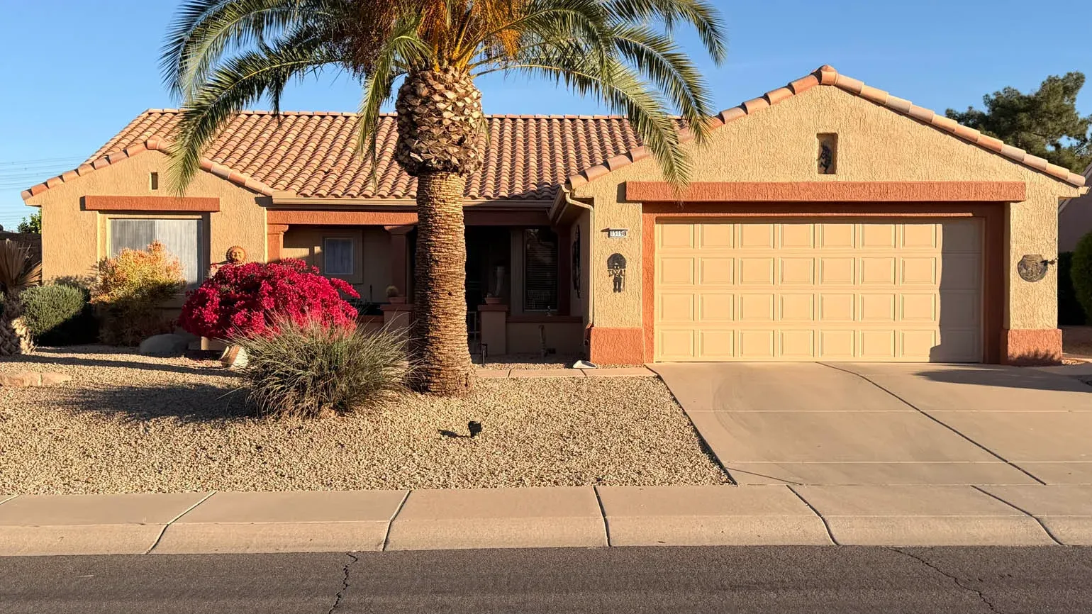 Sun City home with desert landscaping, featuring a beige exterior, red flowering bushes, palm trees, and a two-car garage, illustrating effective exterior painting for summer preparation.